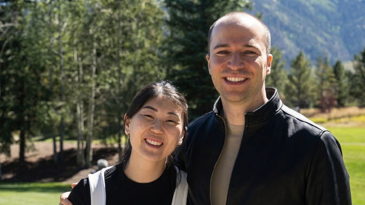 Two people stand together outdoors, smiling, with trees and mountains in the background.