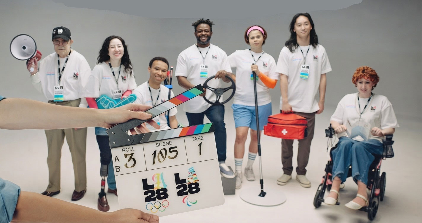 Diverse team of volunteers in white shirts pose in a studio behind a film clapperboard, with a person in a wheelchair on the right.