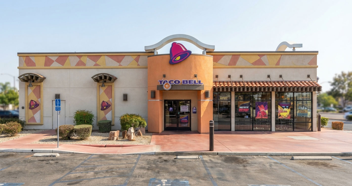 Front view of a Taco Bell restaurant with peach/orange facade, purple bell logo above glass doors, and large windows to the right on a sunny day.