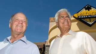 Two smiling older men stand outside a California Pizza Kitchen storefront under a clear blue sky.