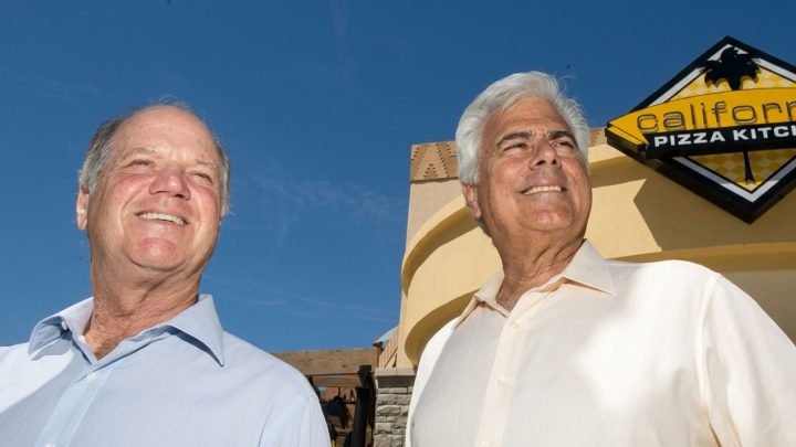 Two smiling older men stand outside a California Pizza Kitchen storefront under a clear blue sky.